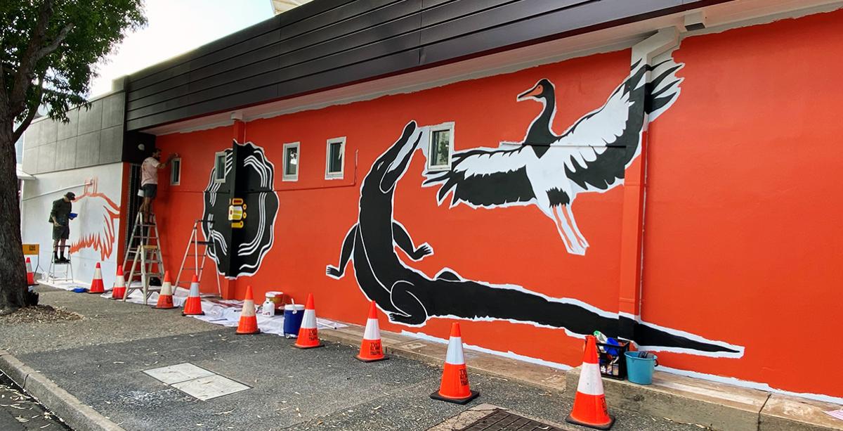 Caleb and Jordan blocking in colour for the Gunimidjina Gwalwa Daraniki, ABC Darwin Wall Larrakia mural, featuring crocodile, magpie goose with orange background